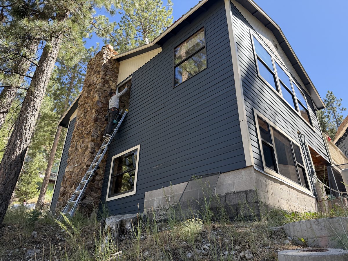 Neon technician on an extension ladder cleaning the upstairs windows of a two-story Mt. Charleston cabin with a stone chimney, surrounded by ponderosa pines
